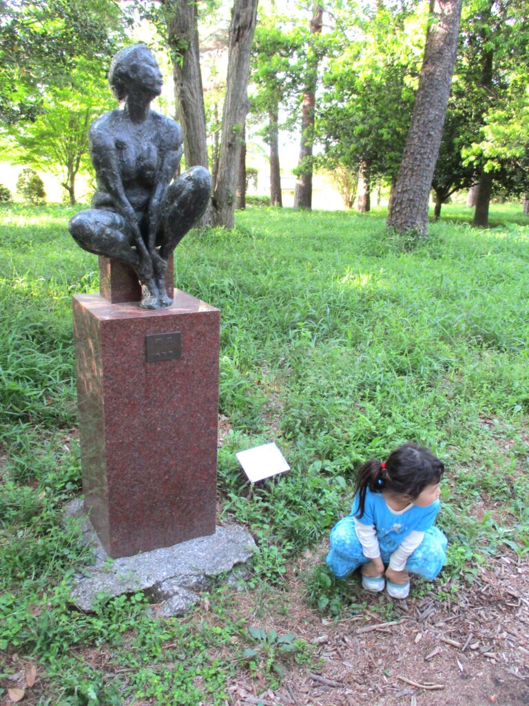 (左)群馬県館林市 彫刻の小径にある木下 繁の「裸婦」(ブロンズ)。右下にキャプションがある。ポーズを真似する娘。